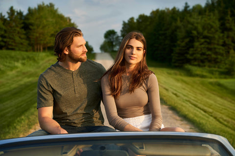 Tall man and woman sitting on back of car along dirt road surrounded by green fields. summer road trip