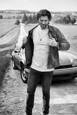 Tall man standing on long dirt road in front of old style car wearing denim jacket and jeans