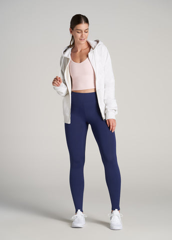 Woman standing wearing dark blue leggings and pale pink tank top layered under white sweater